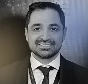 Portrait of a smiling man with dark hair and beard wearing a suit, white shirt, and tie.